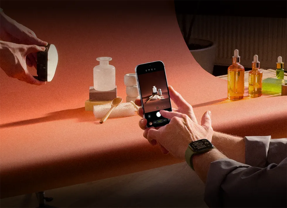 A photoshoot in a workplace setting, one person is using an iPhone to photograph products against a backdrop, one person is holding a light