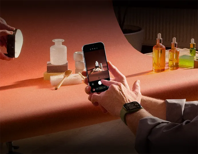 A photoshoot in a workplace setting, one person is using an iPhone to photograph products against a backdrop, one person is holding a light