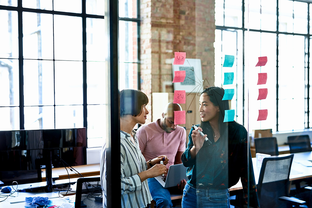 View through glass partition, Hispanic and Chinese businesswomen writing solutions on adhesive notes, smiling and cheerful